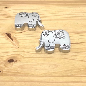 Może przedstawiać: Dwie srebrne figurki słoni z misternymi czarno-białymi wzorami. Słonie stoją na jasnobrązowej drewnianej powierzchni.