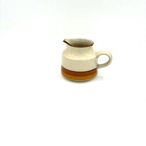 May include: A small, ceramic creamer with a handle and spout. The creamer is off-white with a brown rim and a horizontal band of orange and brown stripes near the base. The creamer is likely vintage or retro-styled.