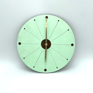 May include: A light green round clock with a wood center and gold hands. The clock face has a sunburst design with small black dots around the edge.