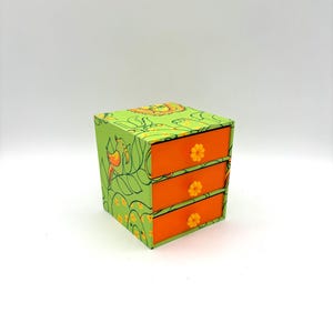 May include: A small, three-drawer jewelry box. The box is painted green with a floral and bird design. The drawers are orange with yellow flower-shaped knobs. The box is a cube shape.