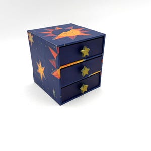May include: A small, navy blue wooden chest of drawers with three drawers. The chest is decorated with a celestial design featuring stars and a sunburst. Each drawer has a gold star-shaped knob.