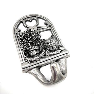 Könnte beinhalten: Ein silberfarbener Metall-Wandhaken mit einer Katze, die aus einem Fenster mit herzförmiger Spitze blickt. Der Fensterrahmen enthält einen Blumentopf mit Blumen. Der Haken ist zum Aufhängen von Gegenständen bestimmt.