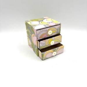 May include: A small three-drawer organiser with a floral pattern in shades of green, yellow, and pink. Each drawer has a white flower knob.