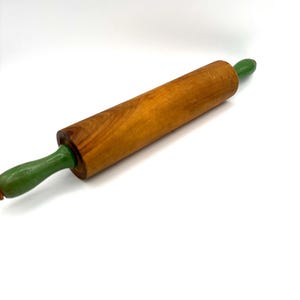 May include: A wooden rolling pin featuring a cylindrical central barrel with a natural light brown wood grain finish and two smooth, rounded handles painted in a solid, vibrant green color, positioned horizontally against a plain white background.