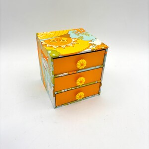 Peut inclure: Une petite commode à trois tiroirs avec un design rétro et psychédélique. Les tiroirs sont orange et ont des boutons en forme de fleurs jaunes. La commode est recouverte d'un motif coloré de soleils, de fleurs et de volutes.
