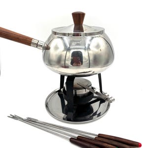 May include: A vintage stainless steel fondue set with a wooden handle and lid knob. The set includes a burner stand, a round base, and several fondue forks with colored tips. The pot has a reflective surface.