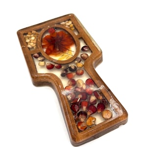 May include: A wooden paddle-shaped decorative item with a clear resin top. The resin encases a red flower, a ladybug, and various seeds. The wooden frame has a natural wood grain finish.