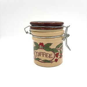 May include: A ceramic coffee canister with a brown lid and a metal latch. The canister has a floral design with red berries and green leaves. The word "COFFEE" is printed in brown letters on the front.