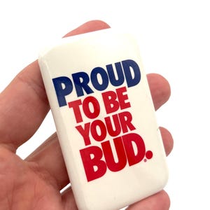 May include: A white rectangular pin with the text "PROUD TO BE YOUR BUD" in blue and red. The pin is held in a hand against a white backdrop.