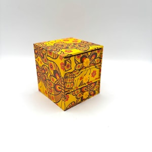 May include: A small, square, three-drawer chest featuring a vibrant yellow background with a repeating pattern of stylized butterflies and small flowers in shades of orange, red, and brown. Each drawer has a small, yellow, flower-shaped knob.