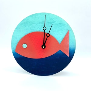 May include: A red fish clock with black hands and a blue and turquoise background. The clock is round and has a white stand.