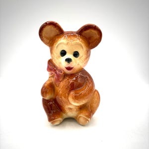 May include: A vintage ceramic bear figurine with a glossy finish. The bear is primarily brown with lighter tan accents on the ears and face. It has black eyes, a red bow, and a smiling red mouth. The figurine is in a sitting position.