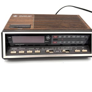 May include: A GE brand AM/FM clock radio with a wood grain top panel, a black front face, and a silver trim border. The device features a digital display, a manual radio tuning dial, and multiple control buttons labeled Reverse, Forward, Sleep, and Wake.