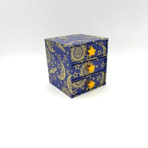 May include: A small three-drawer storage chest featuring a deep blue background with a gold celestial pattern of suns, moons, and stars. Each drawer is equipped with a bright yellow star-shaped knob for opening and closing the compartments.