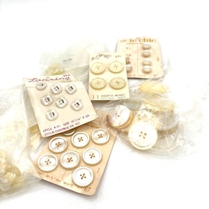 May include: An assortment of vintage buttons in various shapes and sizes. Some are attached to cards with text, including "Le Chic" and "Lansing". The buttons are primarily white and cream-colored, with a few in clear plastic packaging.