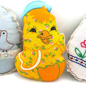 May include: A yellow chick-shaped pillow with a blue hat and orange beak. The chick is decorated with small pink flowers and green leaves. The pillow is part of a set of three decorative pillows.