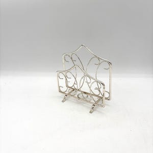 May include: A silver-colored metal letter holder with an openwork design. The holder has a curved top and a decorative, scroll-like pattern. It stands on small feet and is designed to hold mail or documents.