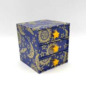 May include: A navy blue, three-drawer jewellery box with a celestial design. It features a gold sun, moon, stars, and UFO pattern. Each drawer has a yellow star-shaped knob. The box is cube-shaped.