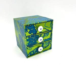 Peut inclure: Une petite boîte en bois à motifs verts et bleus avec quatre tiroirs. Chaque tiroir a une poignée en forme de fleur blanche.