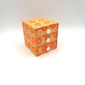 May include: A small, three-drawer chest of drawers with a colourful geometric pattern in shades of orange, yellow, and pink. Each drawer has a white flower-shaped knob.