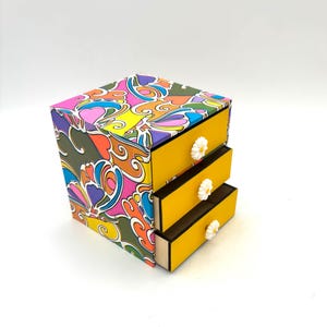 Peut inclure: Une petite commode à trois tiroirs avec un motif psychédélique coloré sur le devant et des tiroirs jaunes avec des boutons en forme de marguerite blanche.