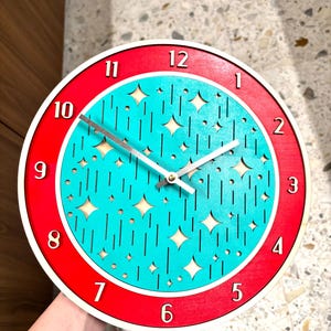 May include: A round wall clock with a red and white frame. The clock face is turquoise with star and line cutouts. The numbers are in a simple, modern font. The clock hands are silver.