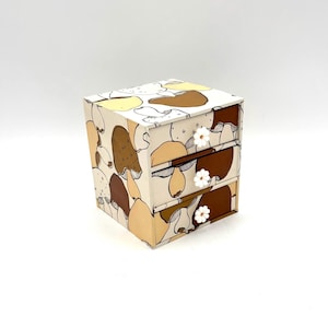 Peut inclure: Petite commode à trois tiroirs ornée d'un motif de champignons fantaisiste dans les tons marron, beige et jaune. La boîte a des boutons de tiroir en forme de fleur blanche. La boîte est de forme cubique.