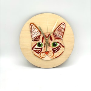 May include: A round wooden clock featuring a detailed, embroidered cat face with green eyes and orange and brown fur. The clock hands are gold-colored, and the clock face is a light wood color.