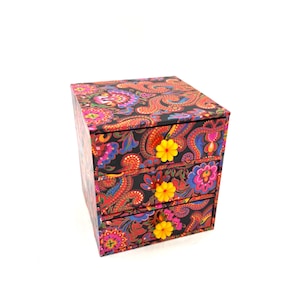 May include: A three-drawer jewellery box with a colourful paisley design in red, blue, pink, and yellow on a black backdrop. Each drawer features a yellow flower. The box is square and rests on a white surface.