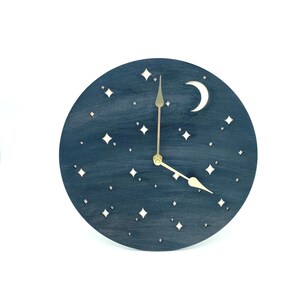May include: A dark blue wooden clock with a crescent moon and stars cut out. The clock has gold hands and a white background.