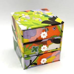 May include: A small, three-drawer box with a floral design. The drawers have white flower-shaped knobs. The box is made of cardboard and has a colorful, abstract pattern.