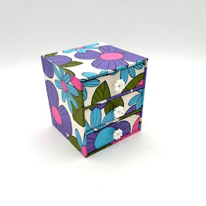 May include: A small square storage chest with three drawers, featuring a white background covered in a repeating pattern of large purple, blue, and pink flowers with green leaves. Each drawer is equipped with a small white flower-shaped knob.