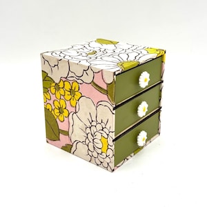 May include: A small, three-drawer dresser with a floral print on the front. The drawers are green and have white daisy-shaped knobs.