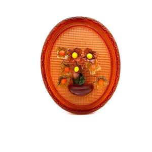 May include: Orange oval-shaped brooch with a raised design of a flower arrangement in a pot. The flowers are orange and yellow with green leaves.