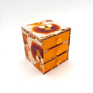 May include: A small, three-drawer wooden storage box. The box has a lion cartoon design on the top and sides, with orange drawers and flower-shaped knobs. The background is white.