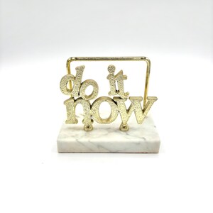 May include: A gold-toned metal desk sign featuring the words "do it now" in a textured, raised font. The metal lettering is attached to a rectangular white marble base with grey veining, supported by a thin gold-toned metal frame structure.