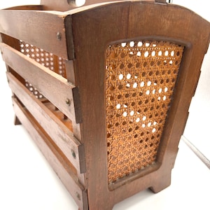 May include: A vintage wooden magazine rack with a woven cane panel. The rack has a handle on the top and is made of dark brown wood.