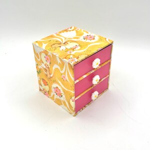 May include: A small, square jewelry box with three drawers. The box is decorated with a yellow floral pattern and has pink drawers with white daisy-shaped knobs. The box is on a white background.