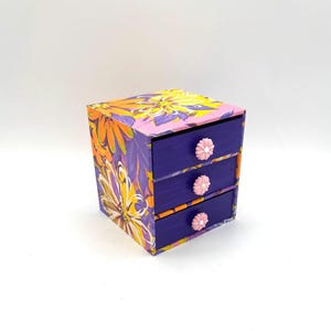 May include: A small wooden box with three drawers. The box is covered in a floral print with yellow, orange, and purple flowers. The drawers are painted a deep purple and have pink flower-shaped knobs.