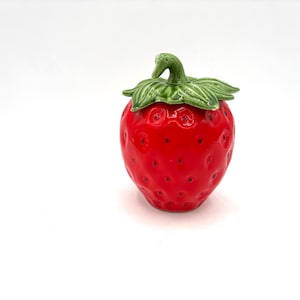 May include: A ceramic strawberry-shaped container with a green stem and leaves. The container is a vibrant red colour with small, textured bumps. The lid is also green, and the container is set against a white background.