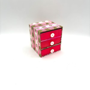 May include: A small, three-drawer chest of drawers with a pink and white floral pattern on the front. The drawers are a bright pink colour with white flower knobs.