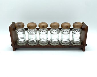 Op de afbeelding: Een vintage kruidenrek met zes heldere glazen potten, elk bekroond met een houten deksel. De potten worden in een donkerbruin houten rek gehouden. De potten hebben een decoratief patroon.