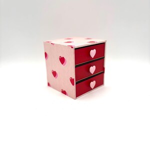 May include: A small pink and red wooden drawer organiser with heart-shaped patterns and pink heart-shaped drawer pulls.