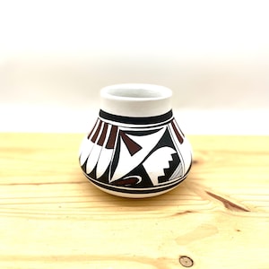 May include: A small, white ceramic pot with a geometric design in black and brown. The pot has a rounded shape and a wide opening.