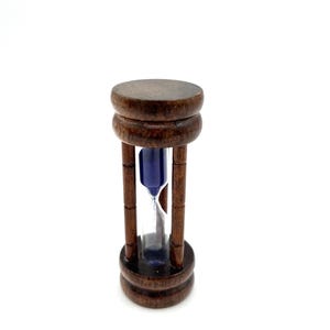 May include: A small hourglass featuring a dark brown wooden frame with a cylindrical top and base. The central glass chamber contains blue sand, which is currently flowing through the narrow neck between the two glass bulbs.