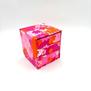 May include: A small square storage chest with three drawers, featuring a vibrant floral pattern in shades of hot pink, light pink, and bright orange. Each drawer is equipped with a small, round, orange flower-shaped knob for opening.