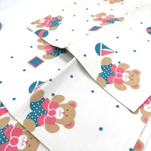 May include: White fabric with a repeating pattern of brown teddy bears wearing blue polka-dot outfits and pink bows. The design includes teal dots and geometric shapes. The fabric is folded, showing multiple layers of the pattern.