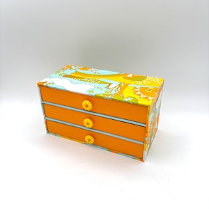 May include: A small, three-drawer storage box with a vibrant floral pattern on the top and sides. The drawers are orange with light blue accents and yellow daisy-shaped knobs. The box is rectangular and sits on a white surface.
