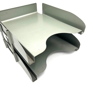 May include: A gray metal desk organizer with three tiers. The organizer has a curved design on the sides and a cutout in the middle of the bottom tier. The metal has a matte finish.
