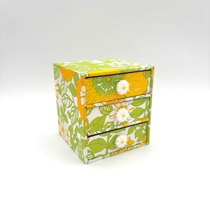May include: A small, three-drawer storage box with a floral pattern in shades of green and yellow. The drawers have white daisy-shaped knobs.